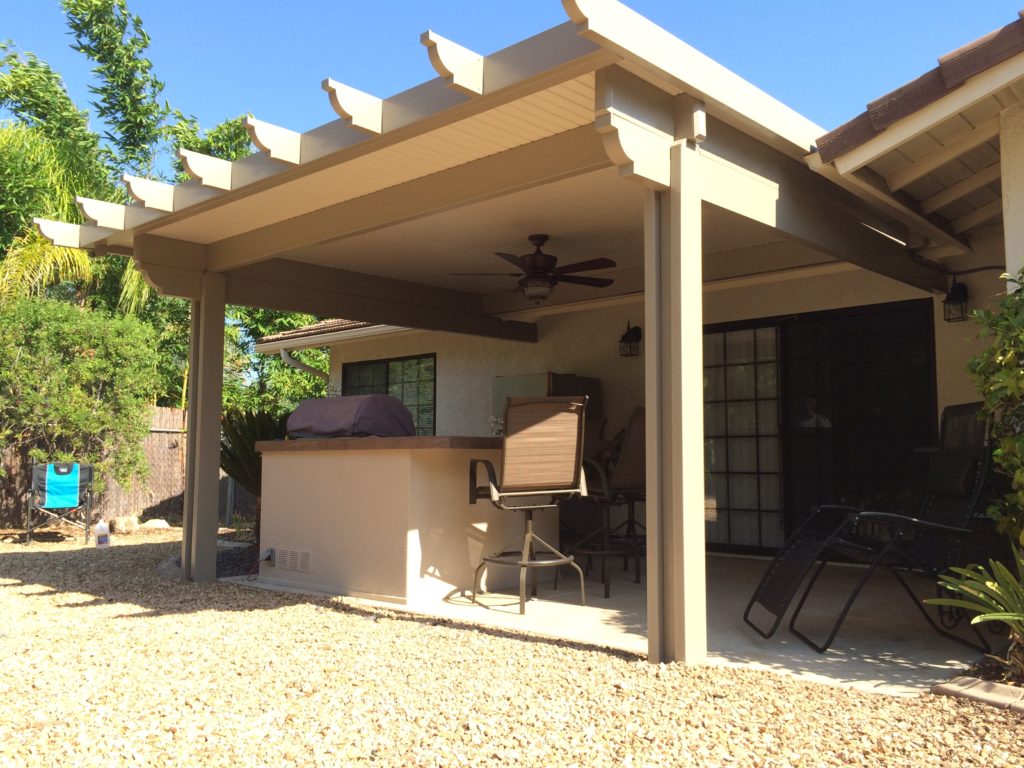 A white solid patio cover over a backyard with a stone fountain and flowering garden beds